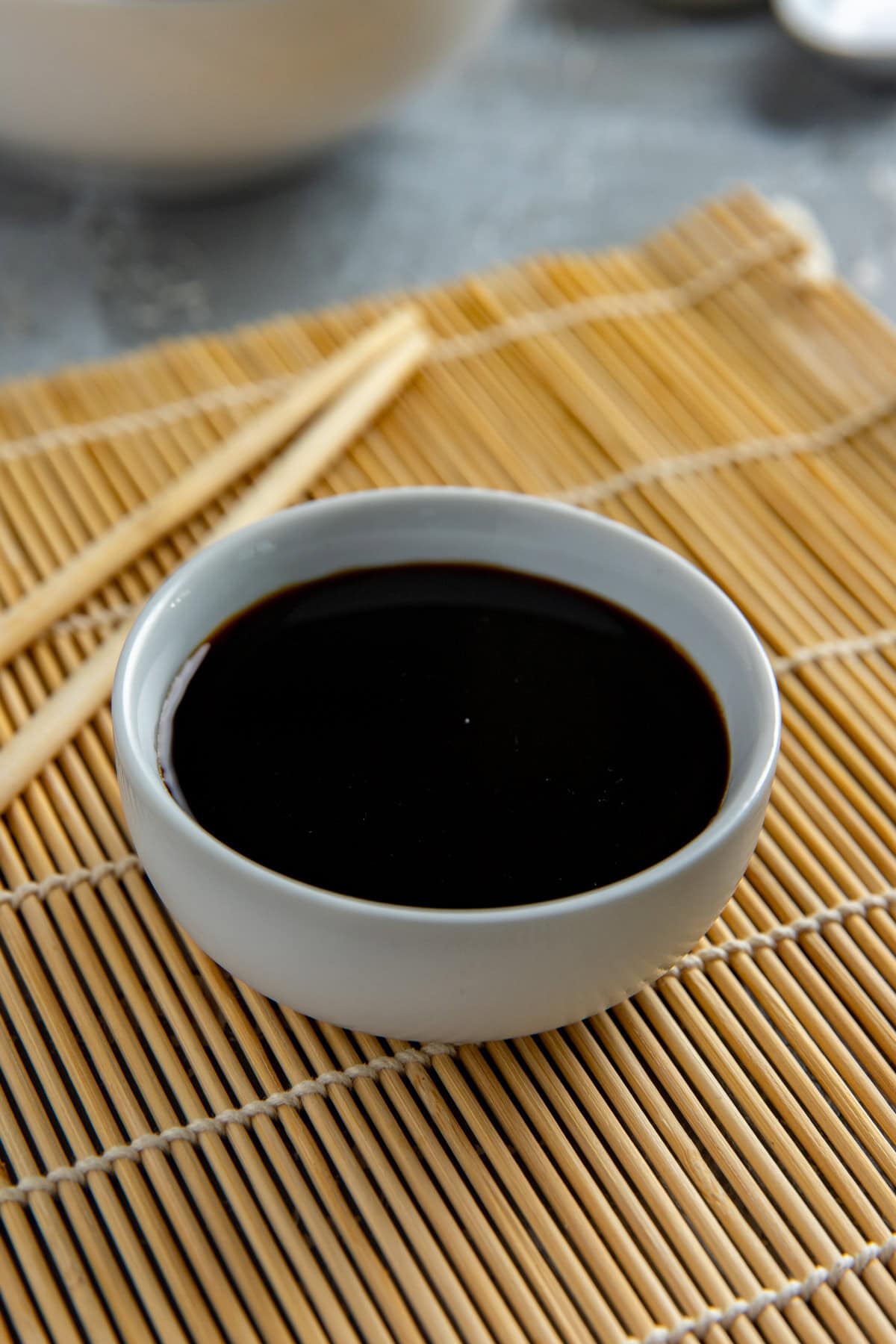 Soy sauce in a small, round white bowl on a wooden place mat with two chop sticks next to the bowl.