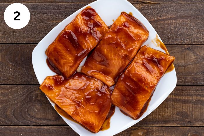 Four uncooked salmon fillets brushed with doctored up BBQ sauce on a white square plate.