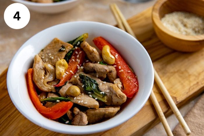 Finished pork stir fry garnished with toasted sesame seeds in a white bowl with chop sticks on the side .