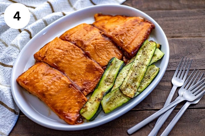 Overhead view of four grilled salmon fillets topped with BBQ sauce on a white platter. Grilled zucchini slices on the side.
