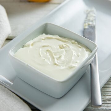 Cream cheese in a square white bowl on a rectangular white platter with a knife on the side. A bagel in the background.