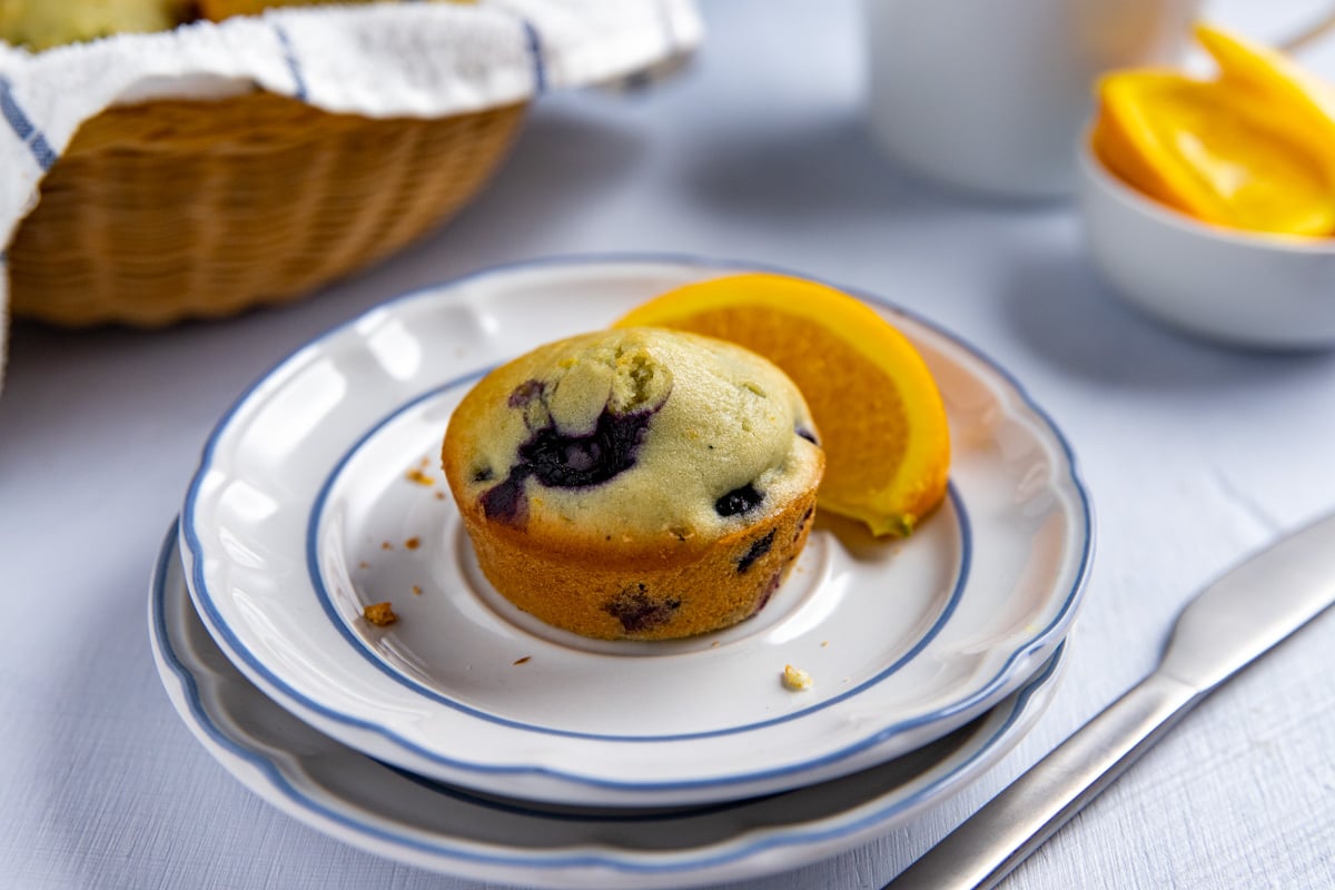 Baked muffin on a white plate with an orange slice on the side. Basket of baked muffins in the background.