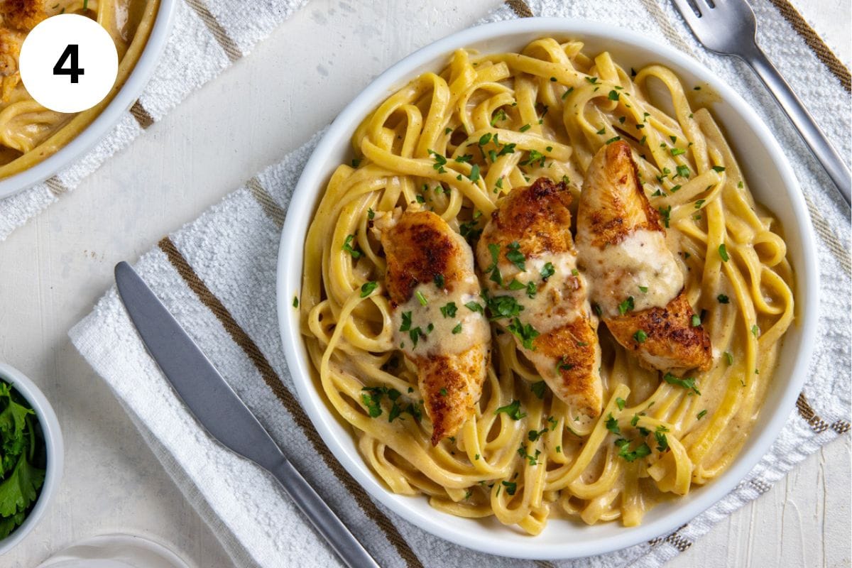 Blackened Chicken Alfredo in a white bowl. Fettucine tossed in alfredo sauce with 3 blackened chicken tenderloins on top. Alfredo sauce drizzled on top of the chicken and fresh parsley sprinkled on top.