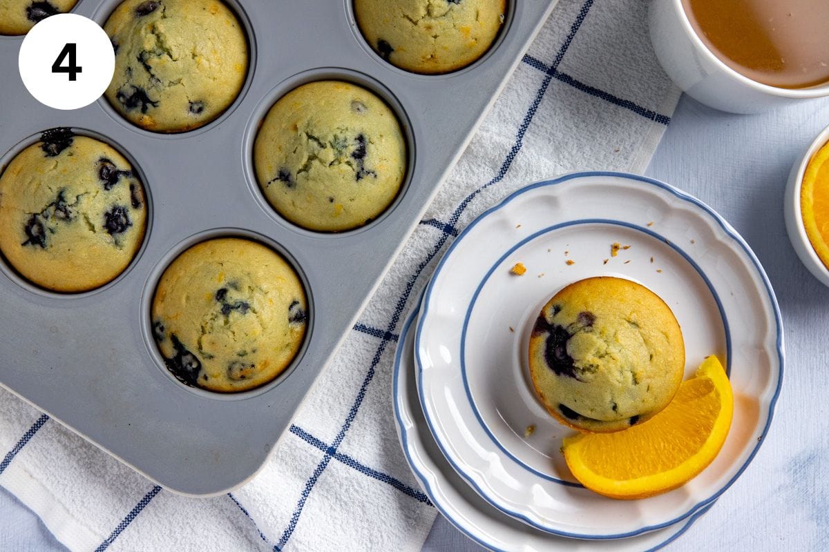 Baked muffin on a white plate with an orange slice on the side. Pan of baked muffins in the background.