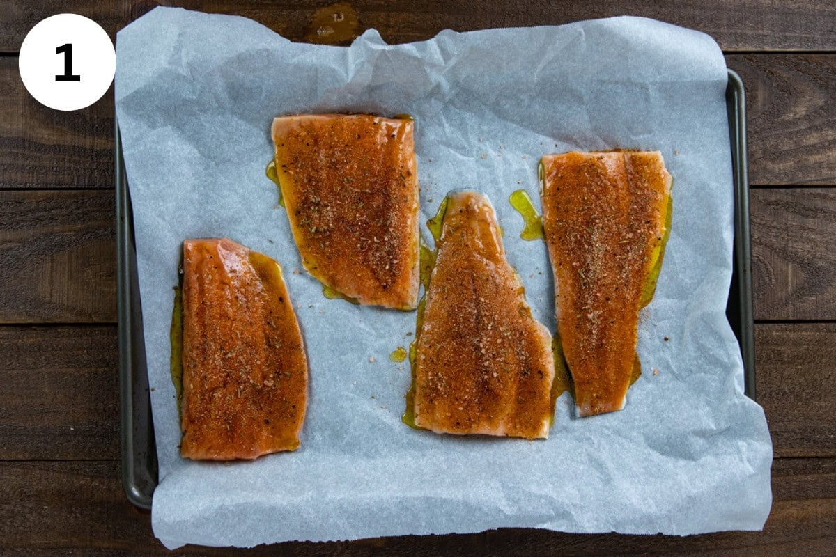 Four uncooked salmon fillets on a parchment lined sheet pan. Topped with honey-olive oil mixture and seasonings.