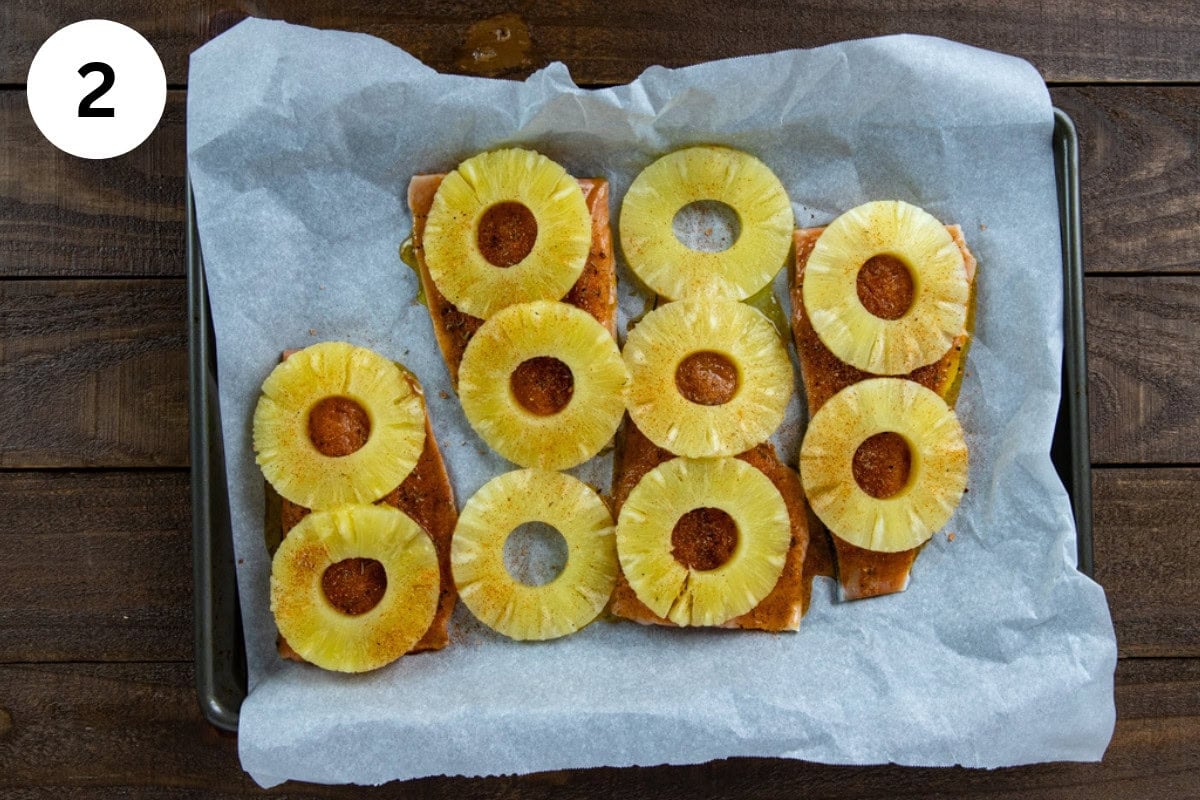 Four uncooked salmon fillets on a parchment lined sheet pan. Topped with honey-olive oil mixture, seasonings, and pineapple rings.