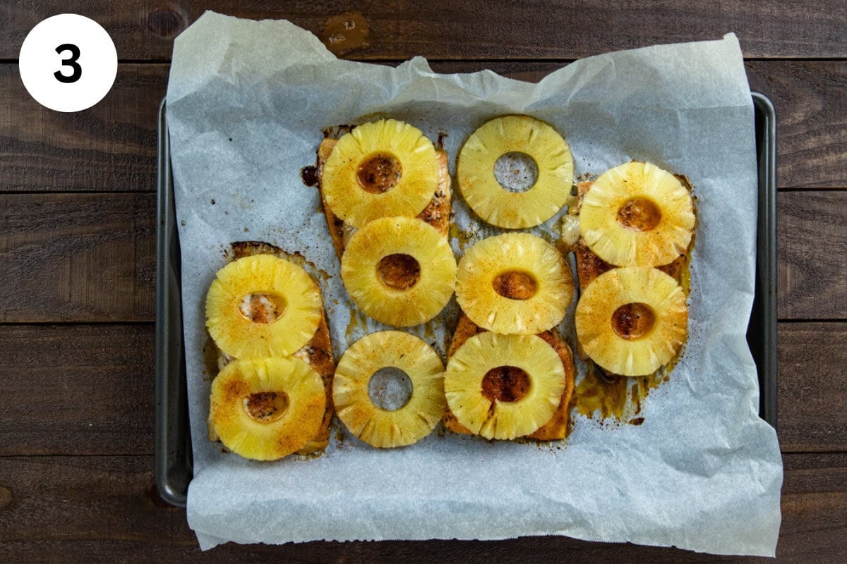 Four baked salmon fillets on a parchment lined sheet pan. Topped with honey-olive oil mixture, seasonings, and pineapple rings.
