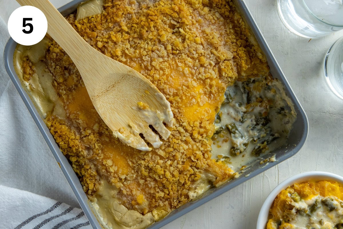 Cooked green bean casserole in square casserole dish with one serving removed. A wooden serving spoon is resting on top of the casserole.