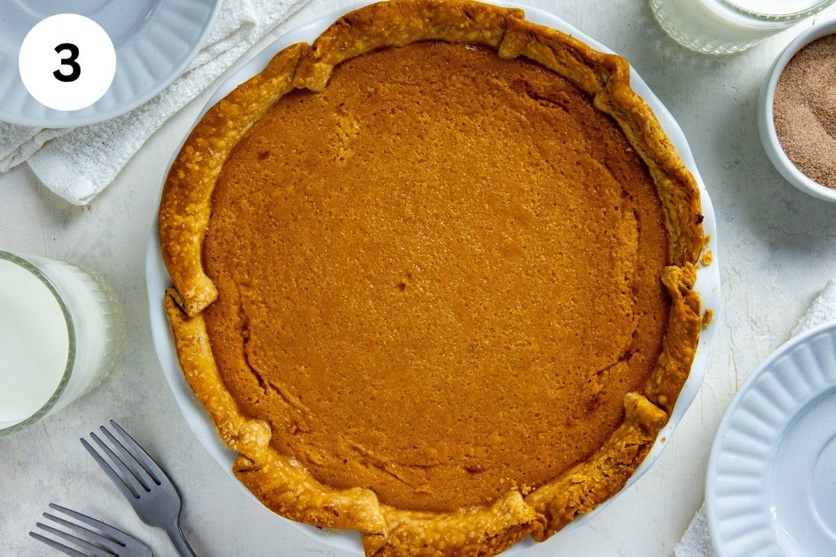 Whole baked sweet potato pie on a white, round plate. Small bowl of cinnamon sugar mixture and glass of milk in the background.