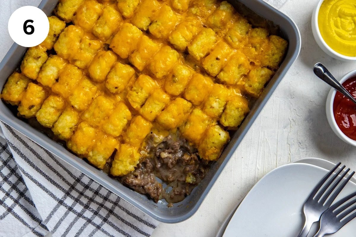 Final baked casserole in the square baking pan with one serving removed. One small container, each containing ketchup and mustard on the side. Two white plates and two forks on the side.